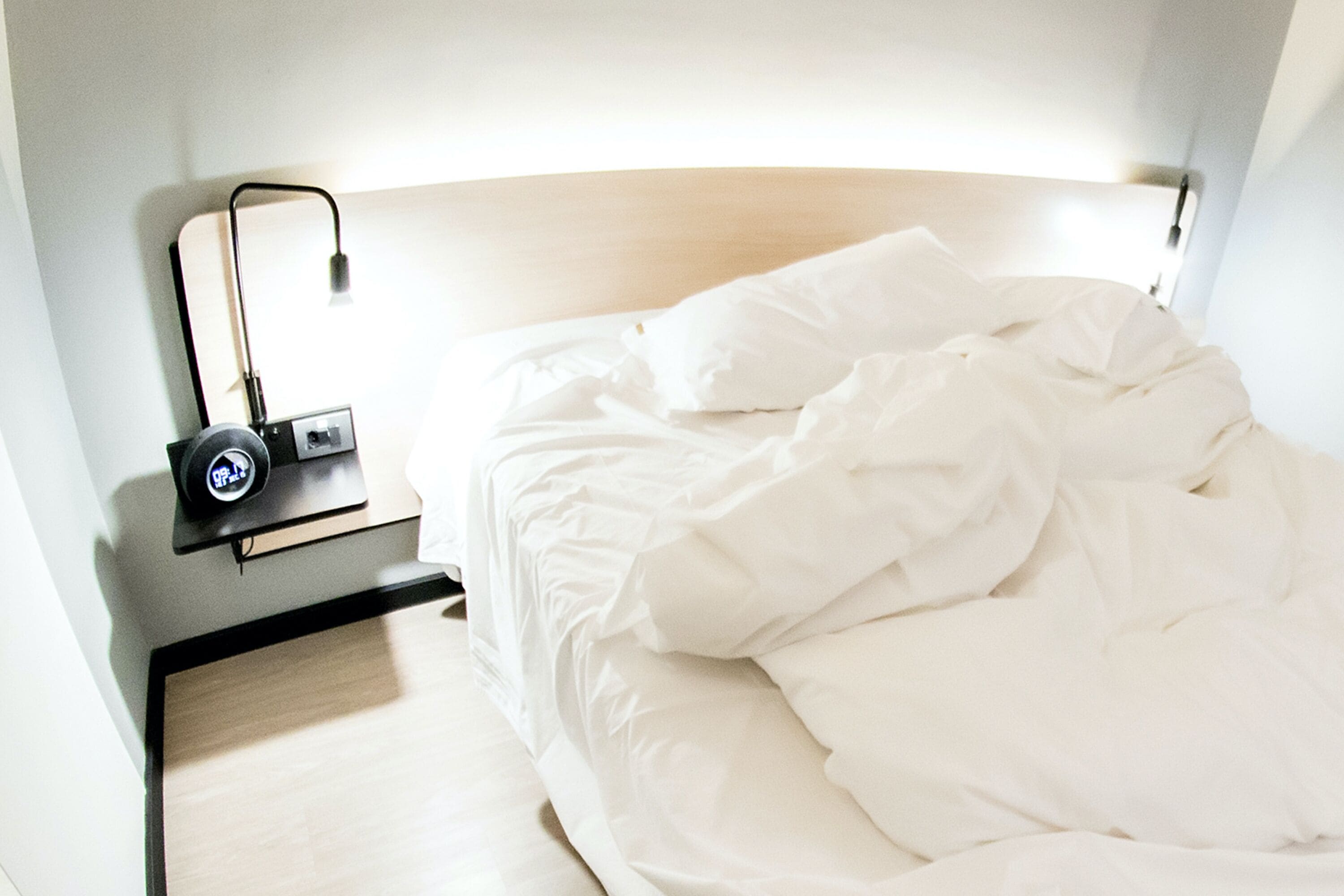 Uma cama bem arrumada com roupa de cama branca, uma mesa de cabeceira com abajur e um relógio fixado na parede.