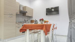 Une cuisine moderne avec des meubles en bois, une table à manger recouverte d’une nappe orange, des chaises blanches et un téléviseur à écran plat fixé au mur.
