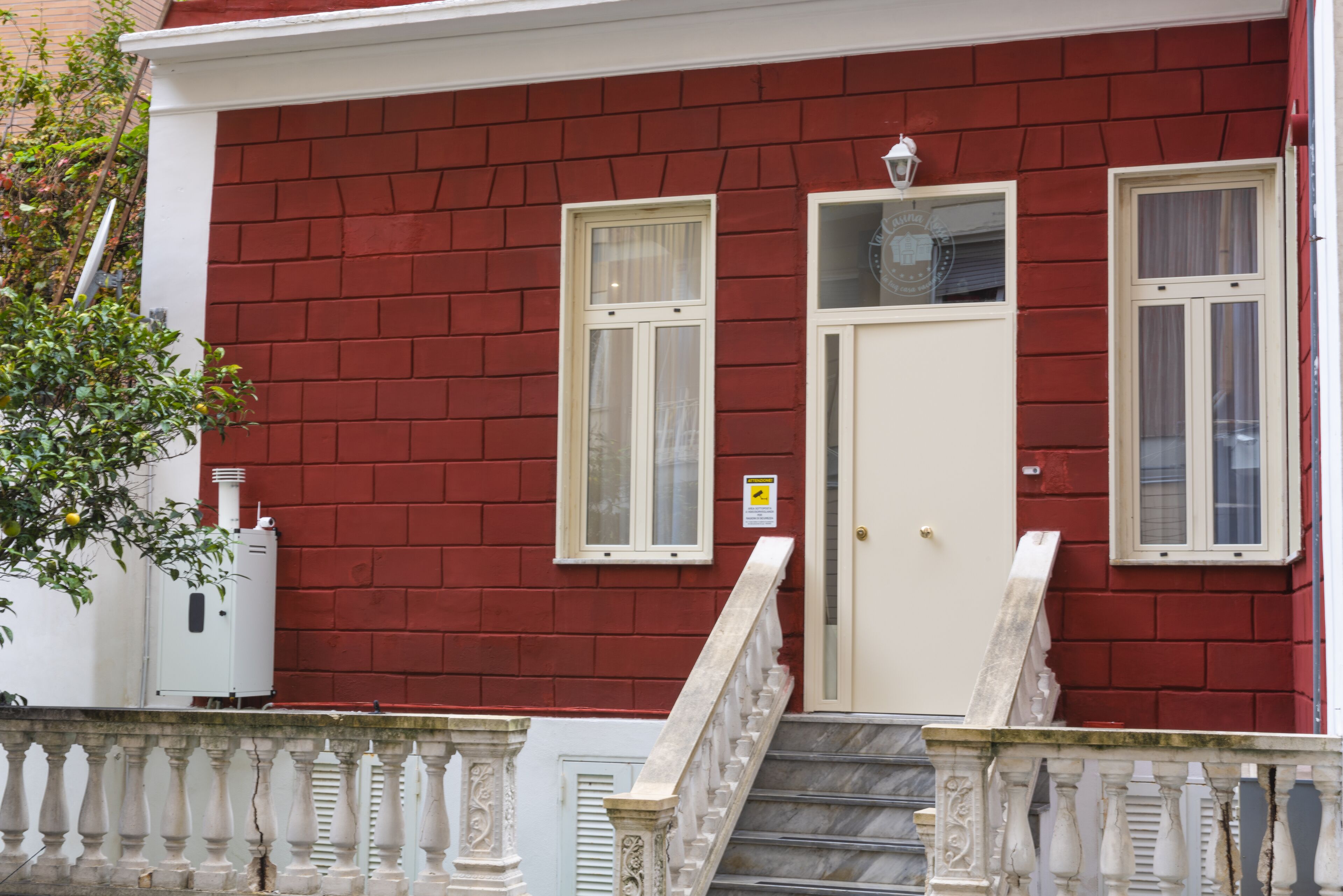 A red brick building with a white door, two windows, and a small balcony with a balustrade.