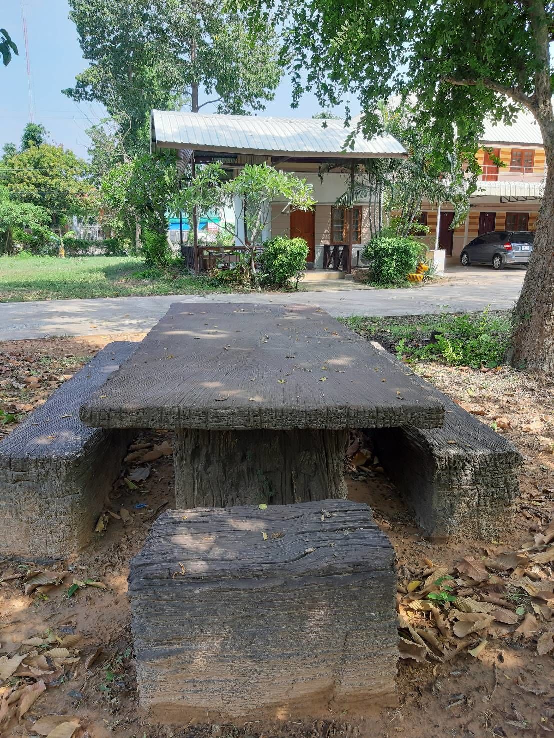 A concrete bench with a rustic design, surrounded by trees and a building with a metal roof in the background.
