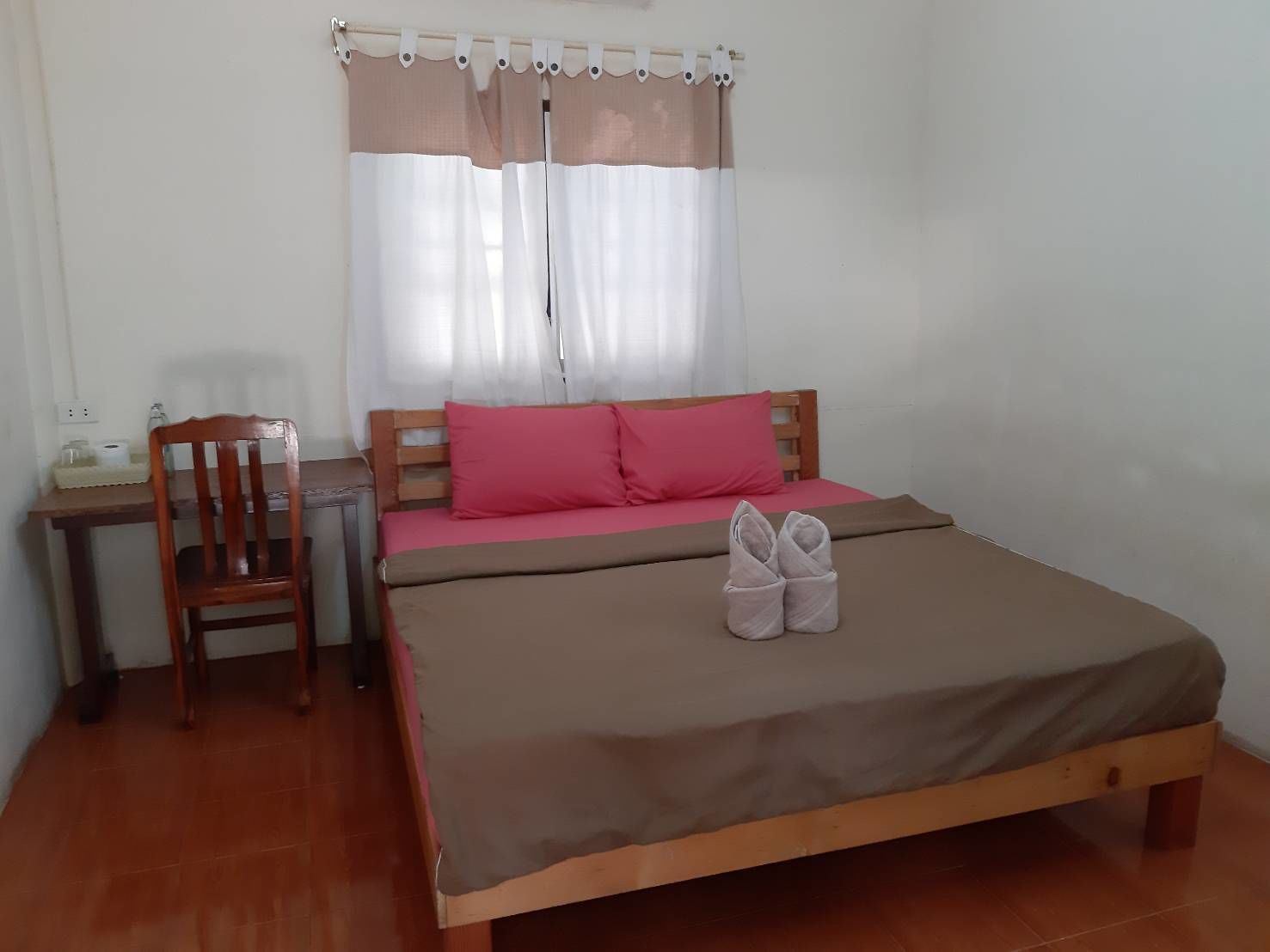 Una cama individual con una colcha marrón, una mesita de noche de madera, una silla y una ventana con cortinas traslúcidas.