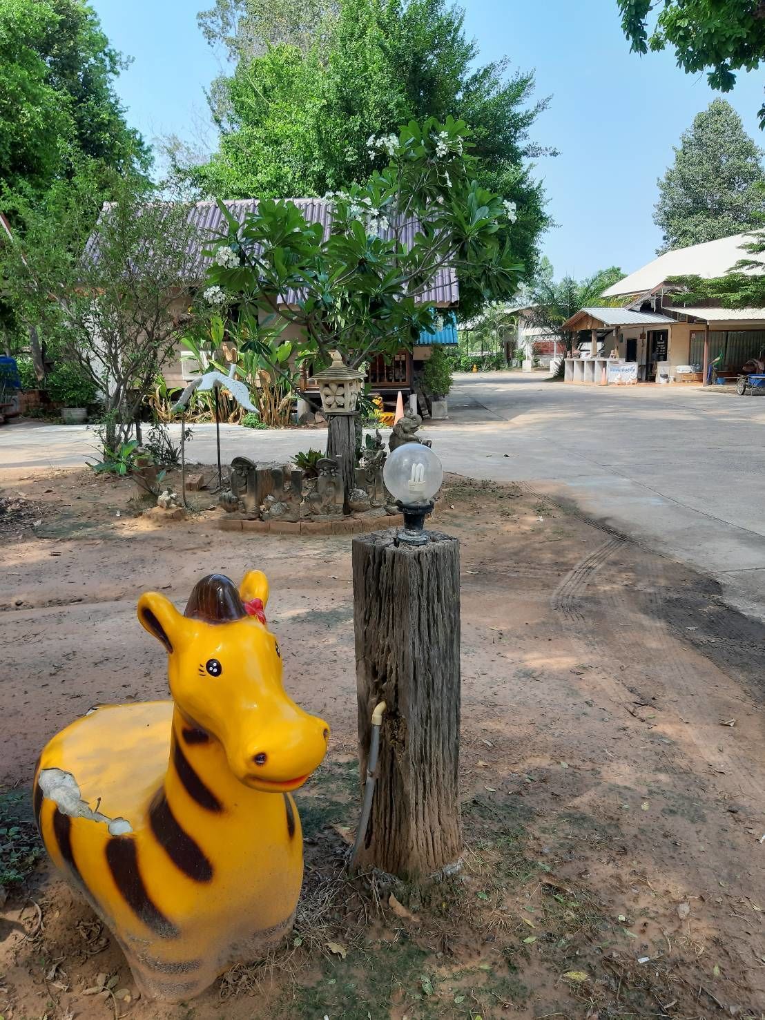 Uma escultura de girafa amarela ao lado de um poste de madeira com um objeto esférico no topo, situada numa área rural com árvores e edifícios ao fundo.