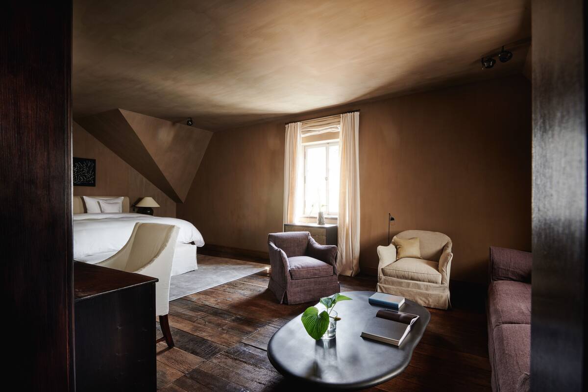 A hotel room with a bed, two armchairs, a small table with a plant, and a window with curtains.
