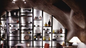 A cozy dining area with a round wooden table, cushioned chairs, and a wine glass. A bookshelf filled with bottles and decorative items is in the background. - Hotel PURS (Andernach)