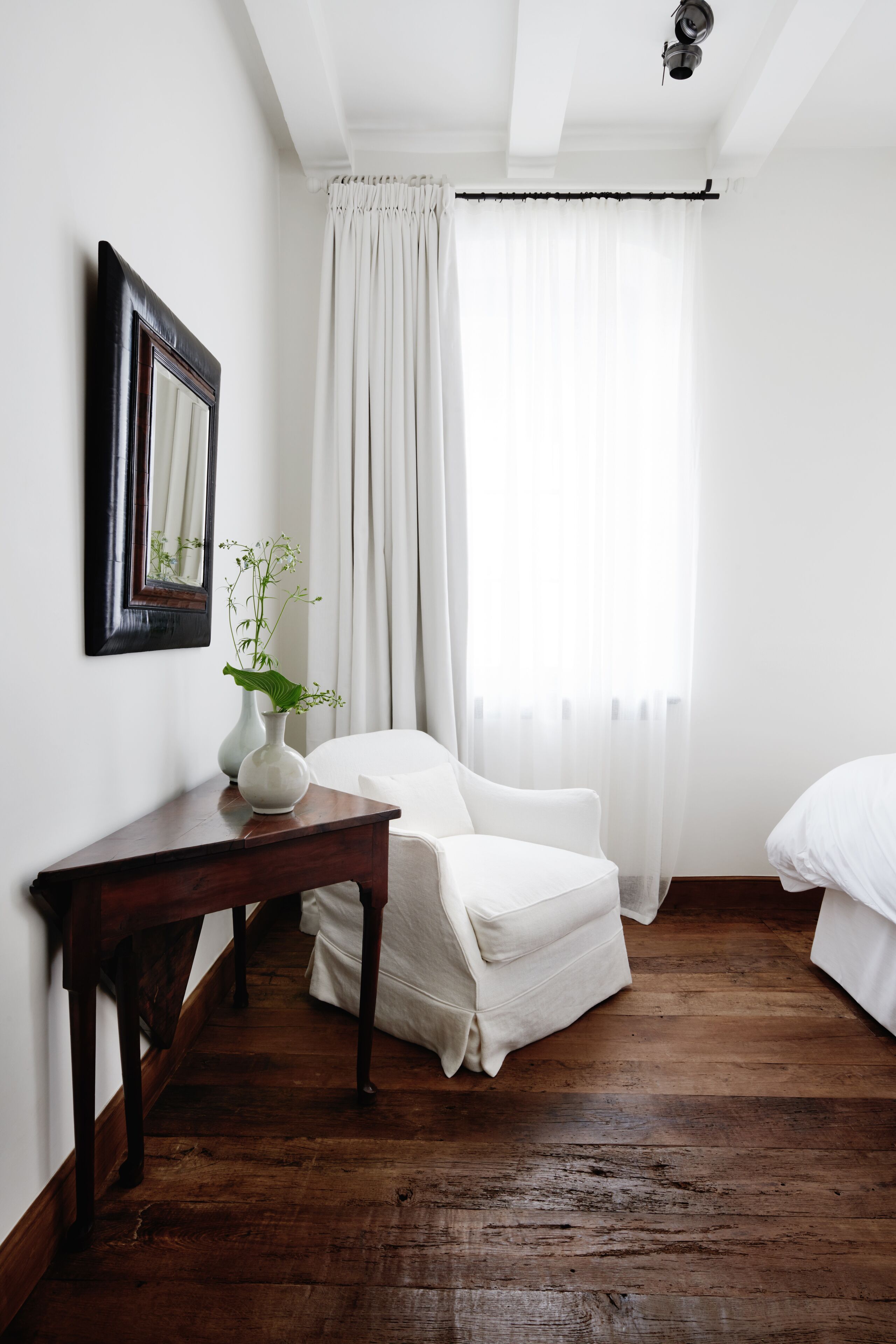 A room with a wooden floor, a white chair, a small table with a vase, and a bed.