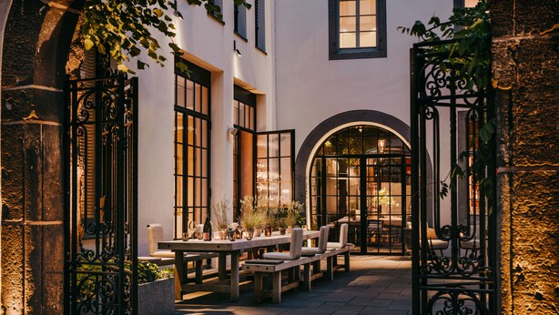 An outdoor patio area with a wooden table and benches, surrounded by white walls and large windows. - Hotel PURS (Andernach)