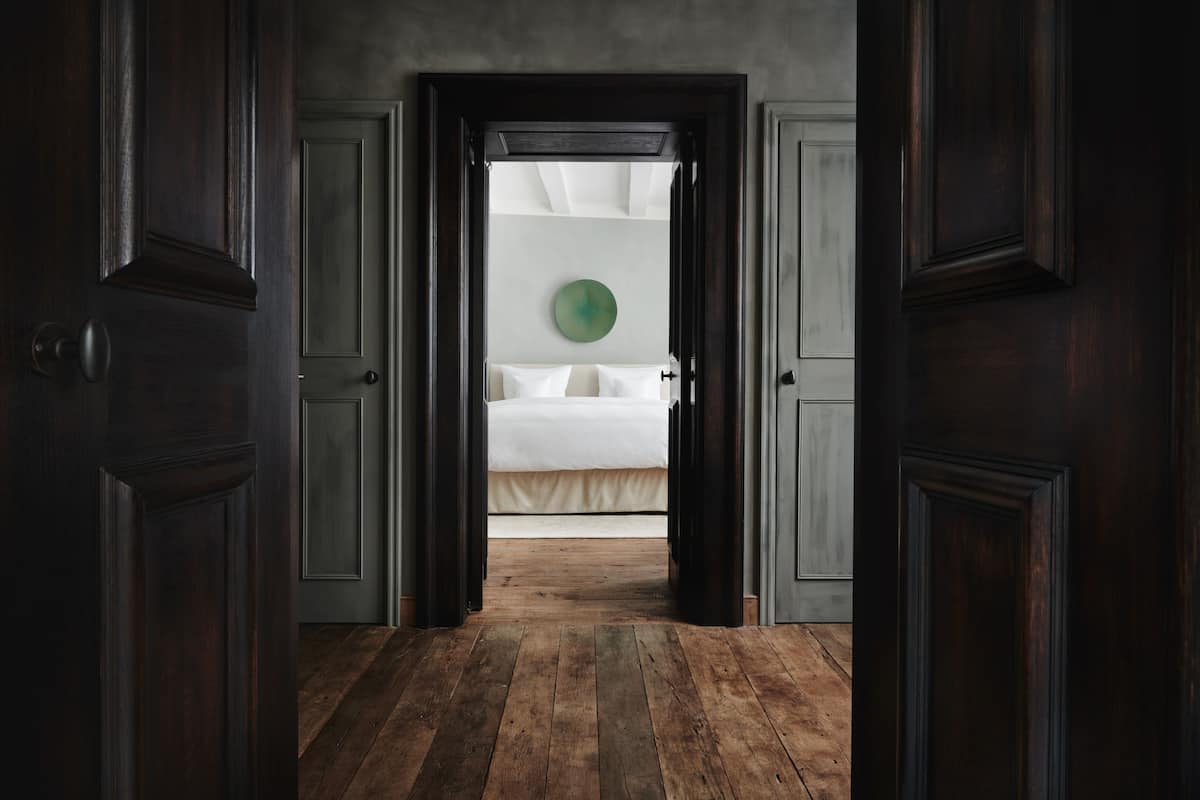 A wooden-floored hallway leading to a bedroom with a bed, a circular wall decoration, and a door on the left.