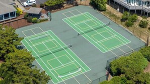 An aerial view of a tennis court complex with multiple courts, surrounded by trees and buildings. - The Myllet at Ships Watch in Duck (Duck)