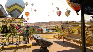 A rooftop terrace with a view of the desert, featuring a cushioned chair, potted plants, and a variety of colorful hot air balloons in the background. - Goreme Reva Hotel (Nevsehir)