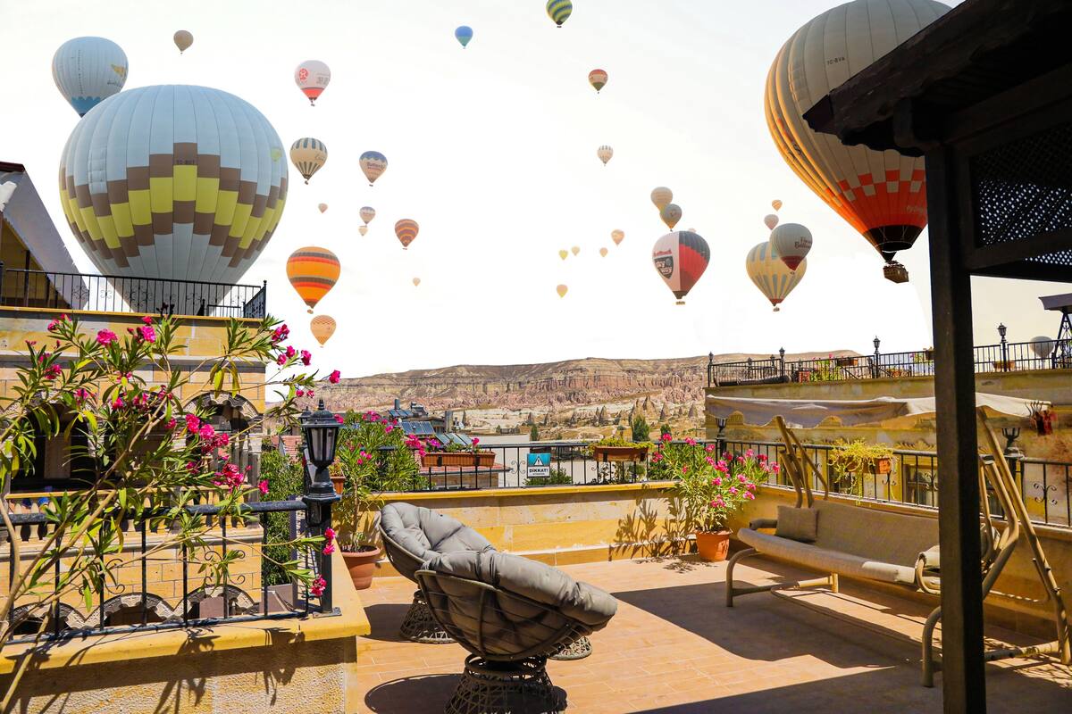 A rooftop terrace with a view of the desert, featuring a cushioned chair, potted plants, and a variety of colorful hot air balloons in the background.
