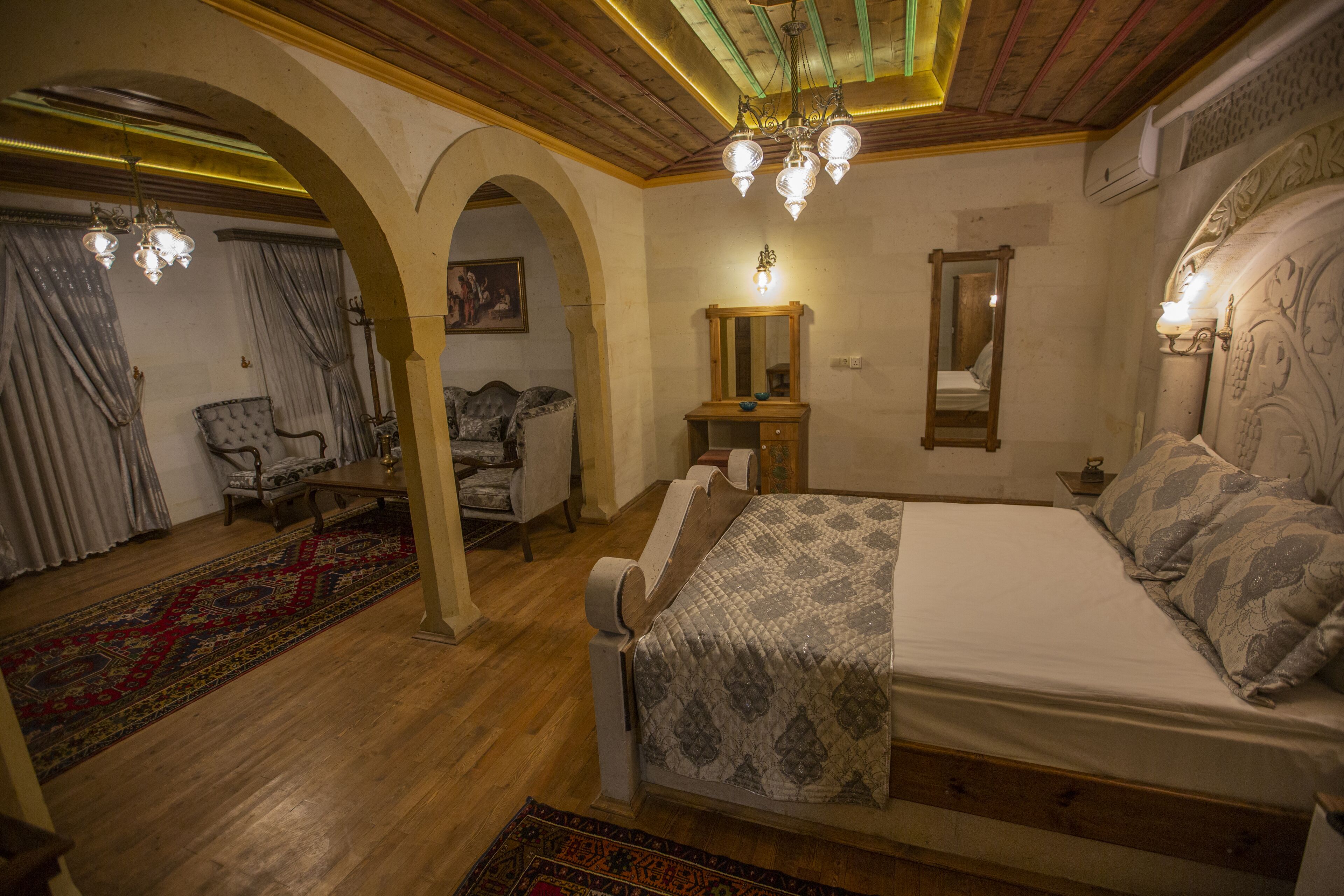 A bedroom with a large bed, wooden floor, arched doorway, chandelier, and a mirror on the wall.
