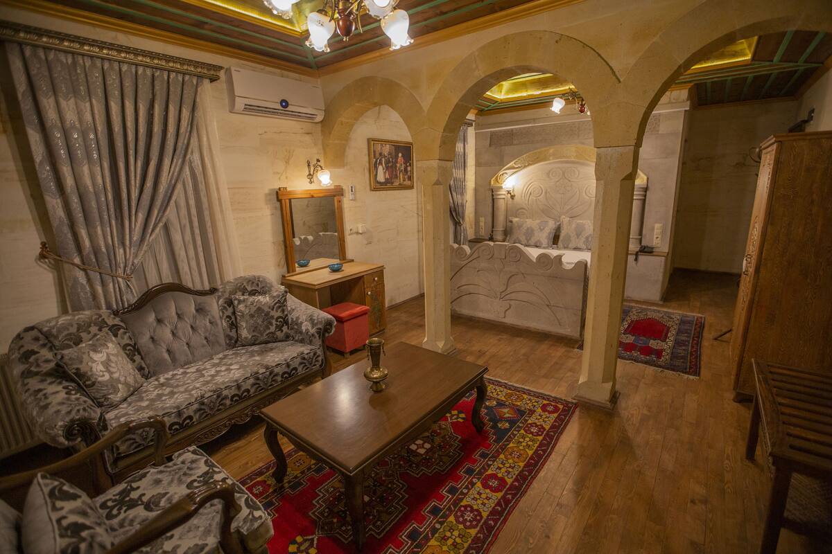 A room with a sofa, a wooden table, and a bed with a headboard. There is a mirror on the wall and a red rug on the floor.