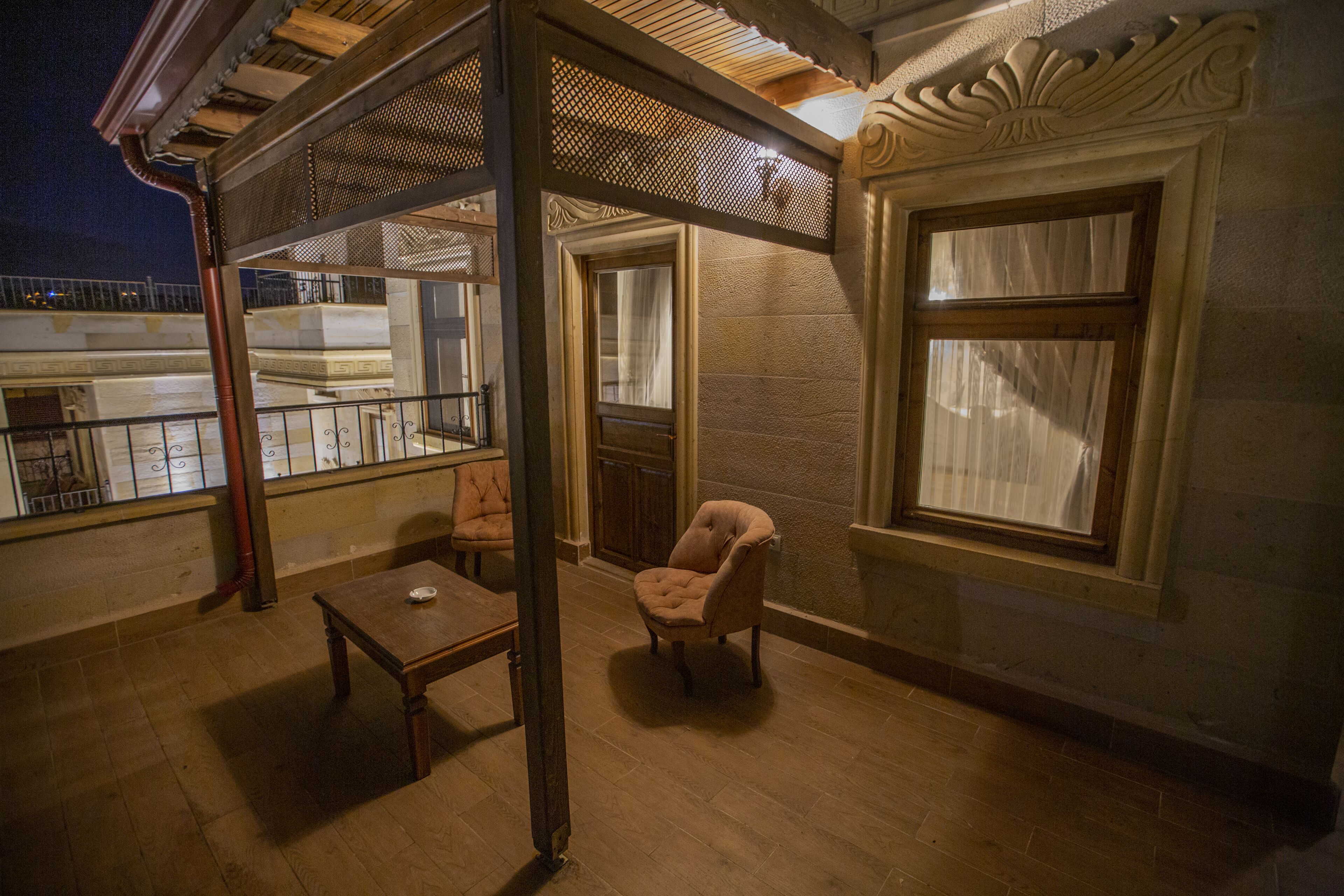 A balcony with a wooden table and a cushioned chair, a window with curtains, and a view of the building's exterior.