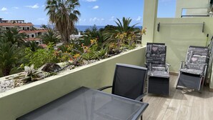 A balcony with a table and two chairs, a view of the ocean, and a variety of plants and trees.