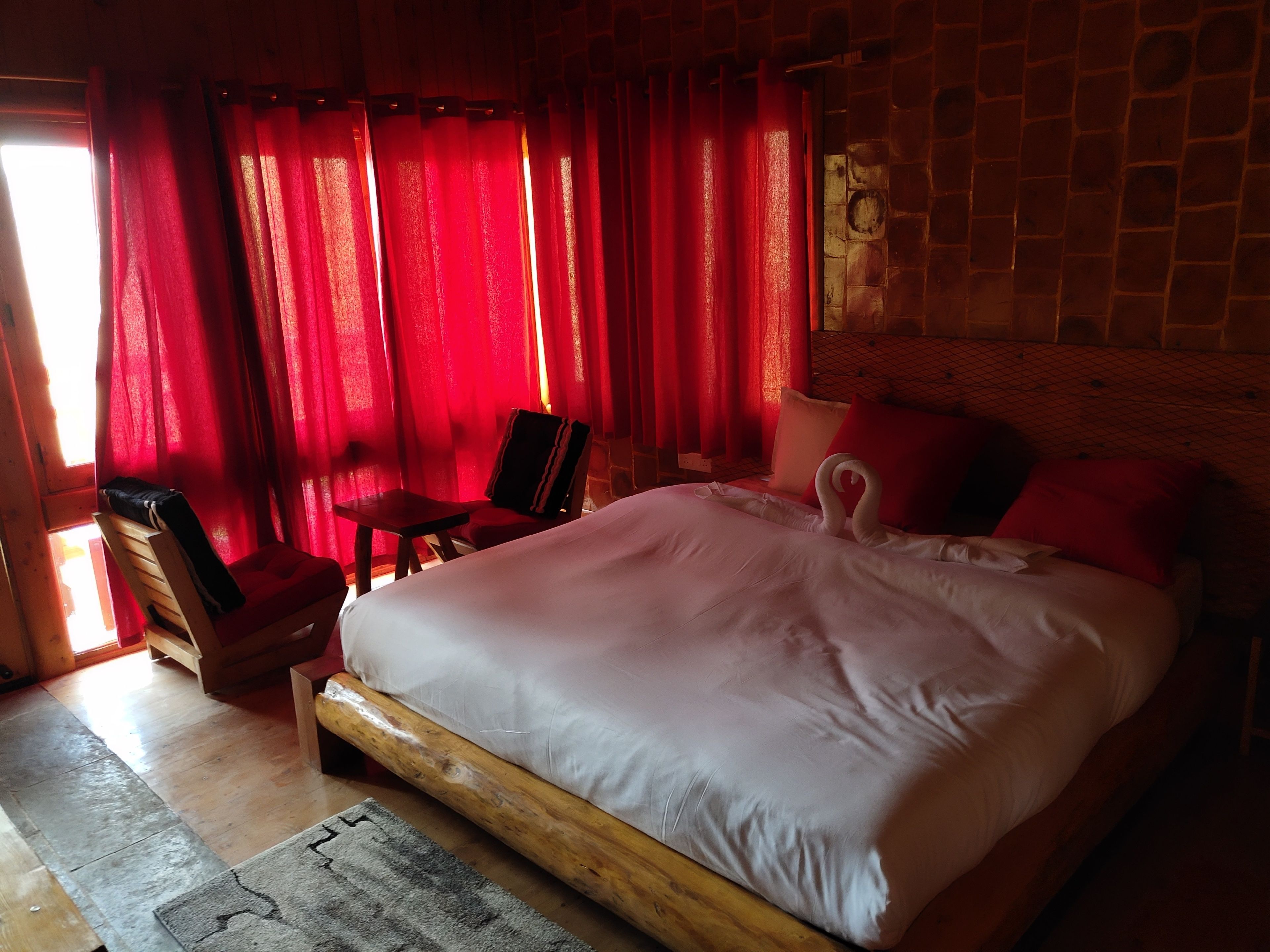 Een kamer met een groot bed, een houten stoel, een klein tafeltje en rode gordijnen.