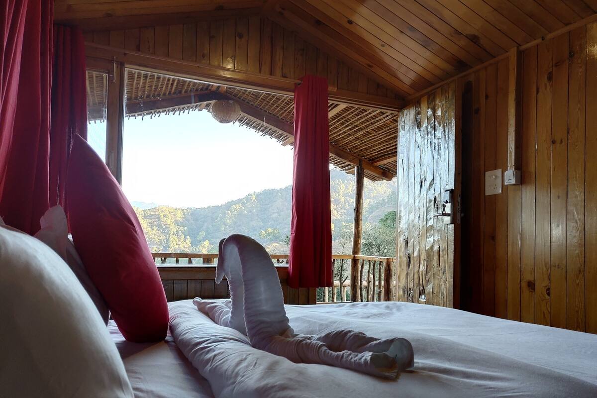 A wooden cabin room with a bed, red curtains, and a view of mountains.