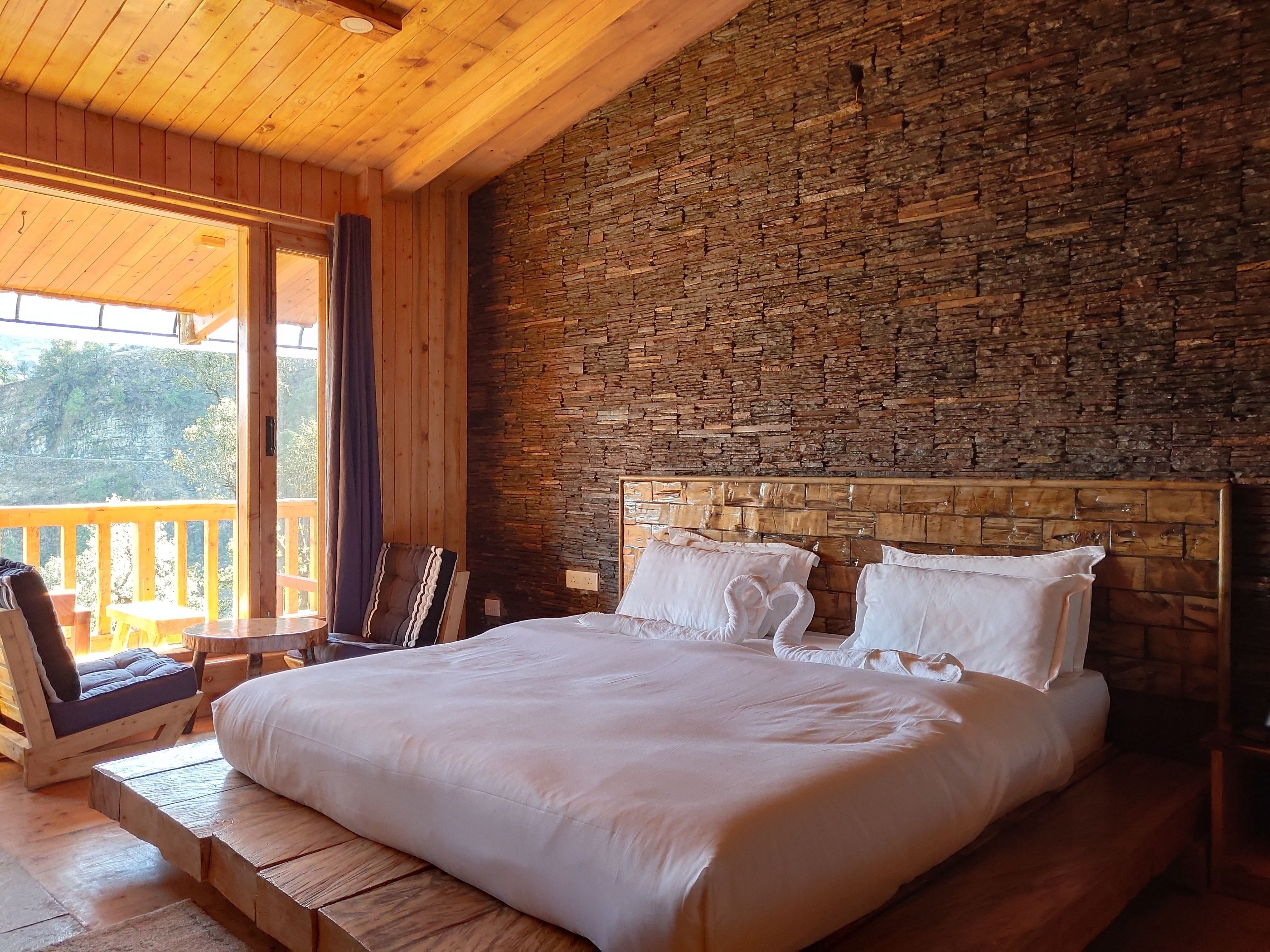 Ein Schlafzimmer einer Holzhütte mit einer Steinwand, einem großen Bett mit weißer Bettwäsche, einer hölzernen Bank und Blick ins Freie.