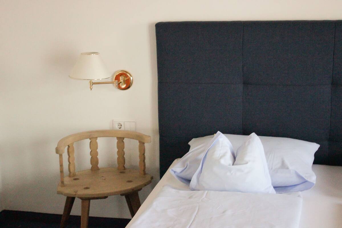 Un lit bien fait, avec du linge de lit blanc et des oreillers, une chaise en bois et une lampe fixée au mur.