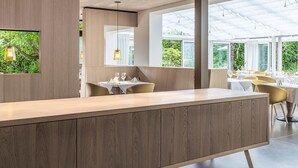 A modern restaurant with a wooden counter, yellow chairs, and large windows offering a view of greenery. - Design-Hotel Ballguthof (Lana)