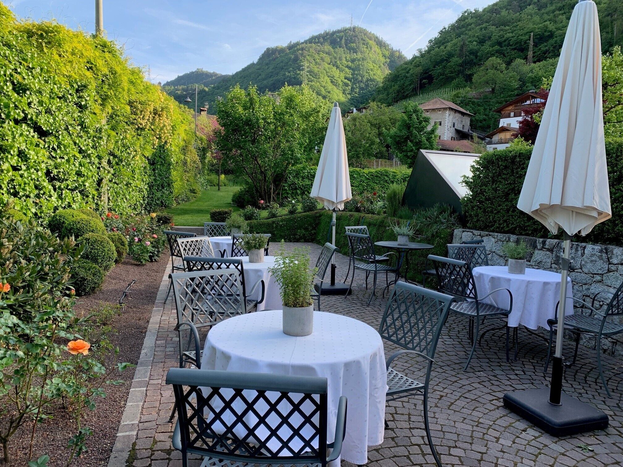 An outdoor patio with round tables and chairs, surrounded by lush greenery and a mountain backdrop.