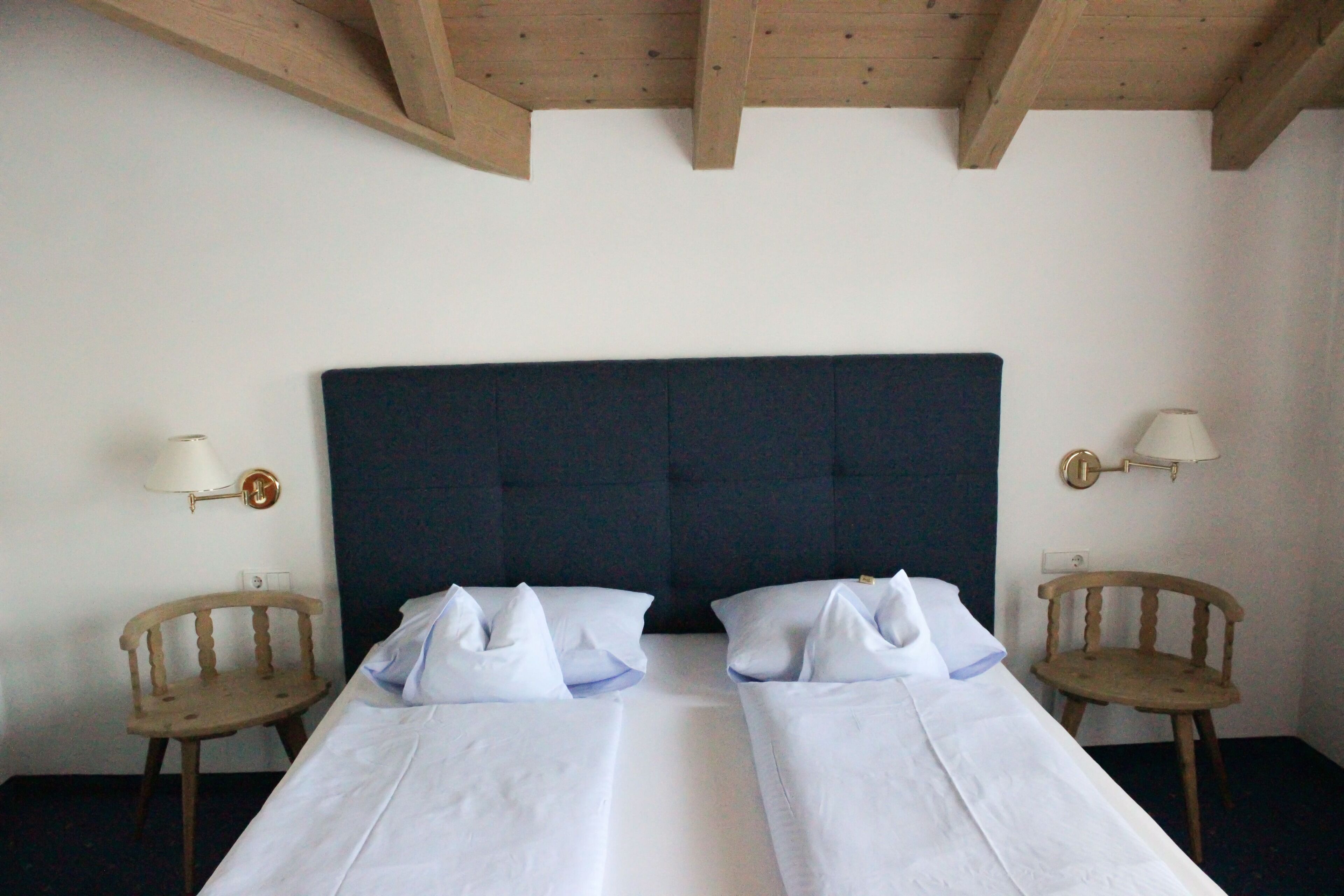 Una habitación de hotel con dos camas individuales, sillas de madera, un cabecero oscuro y lámparas fijadas a la pared.