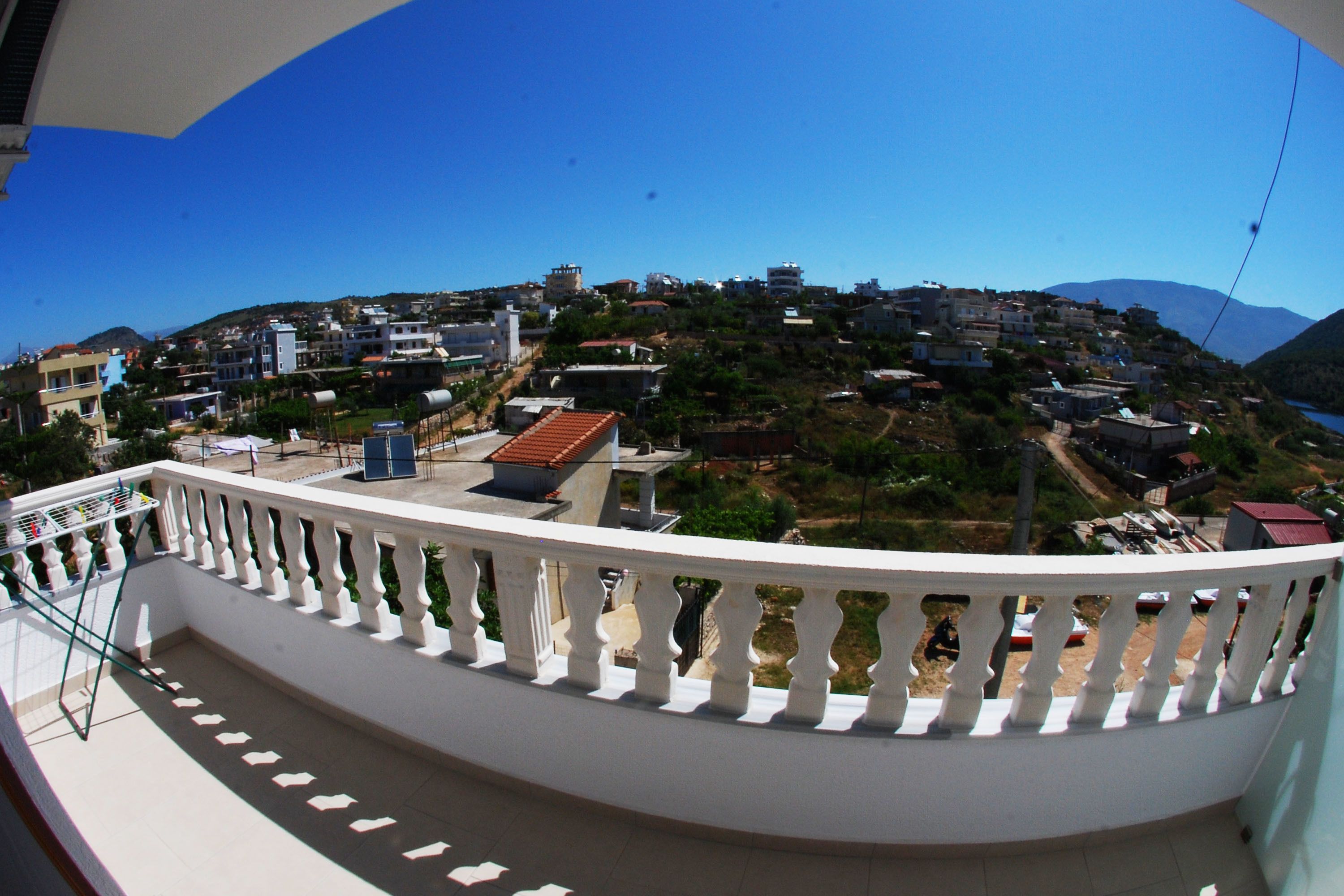 Un balcón con barandilla blanca que da a una villa en la ladera, con edificios y un cielo azul despejado.