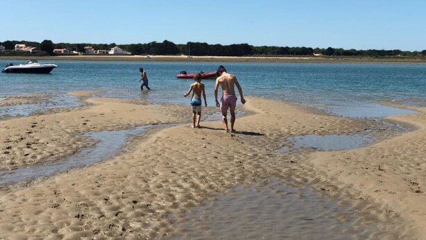 Una playa con gente caminando, un barco en el agua y casas al fondo.