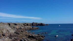 Una costa rocosa con una estructura de piedra histórica, mar azul cristalino y un pequeño bote.