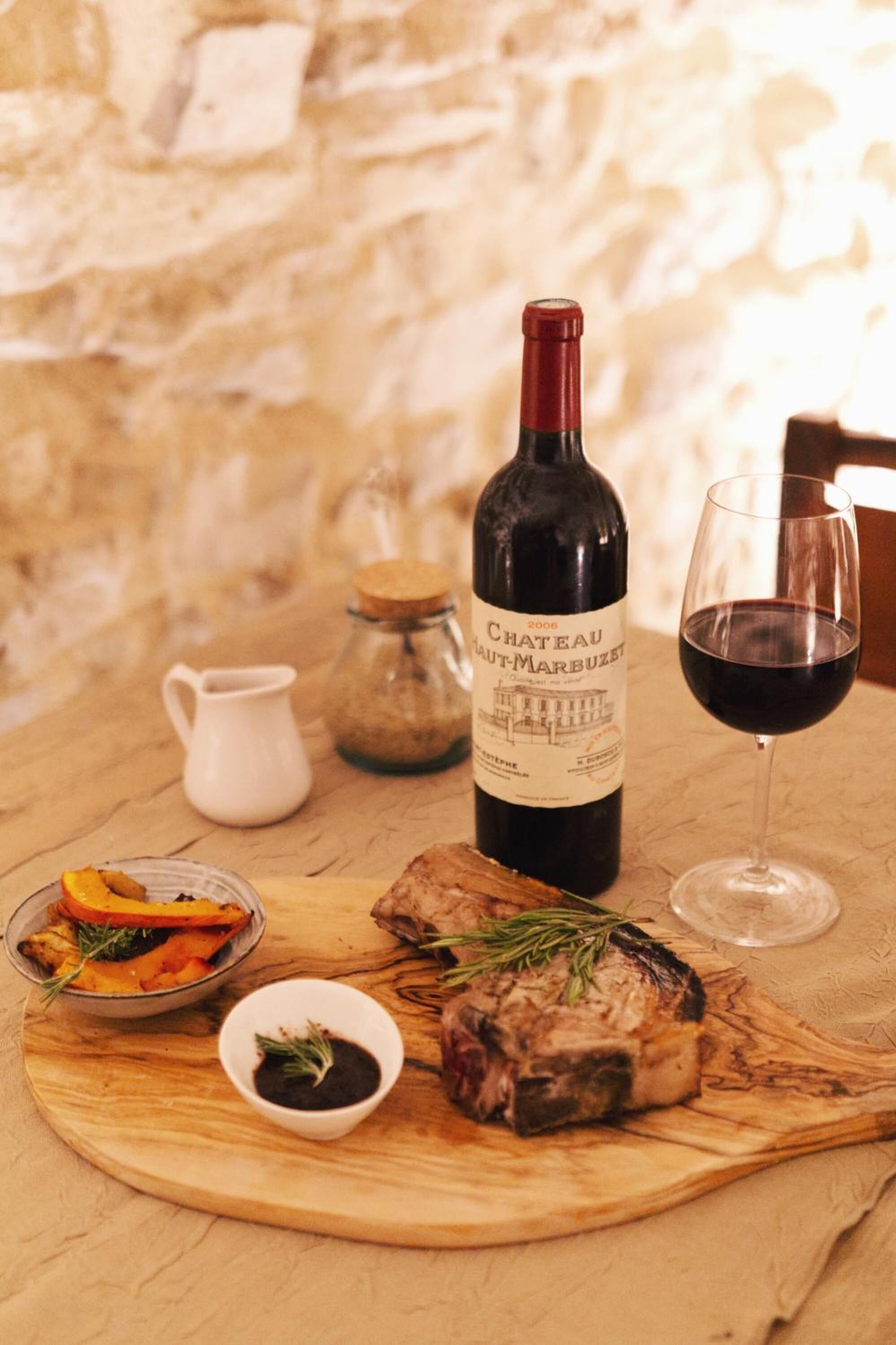Une bouteille de vin Château Haut-Marblé, un verre de vin rouge, de la viande rôtie et un plat de légumes rôtis, le tout présenté sur une planche en bois.