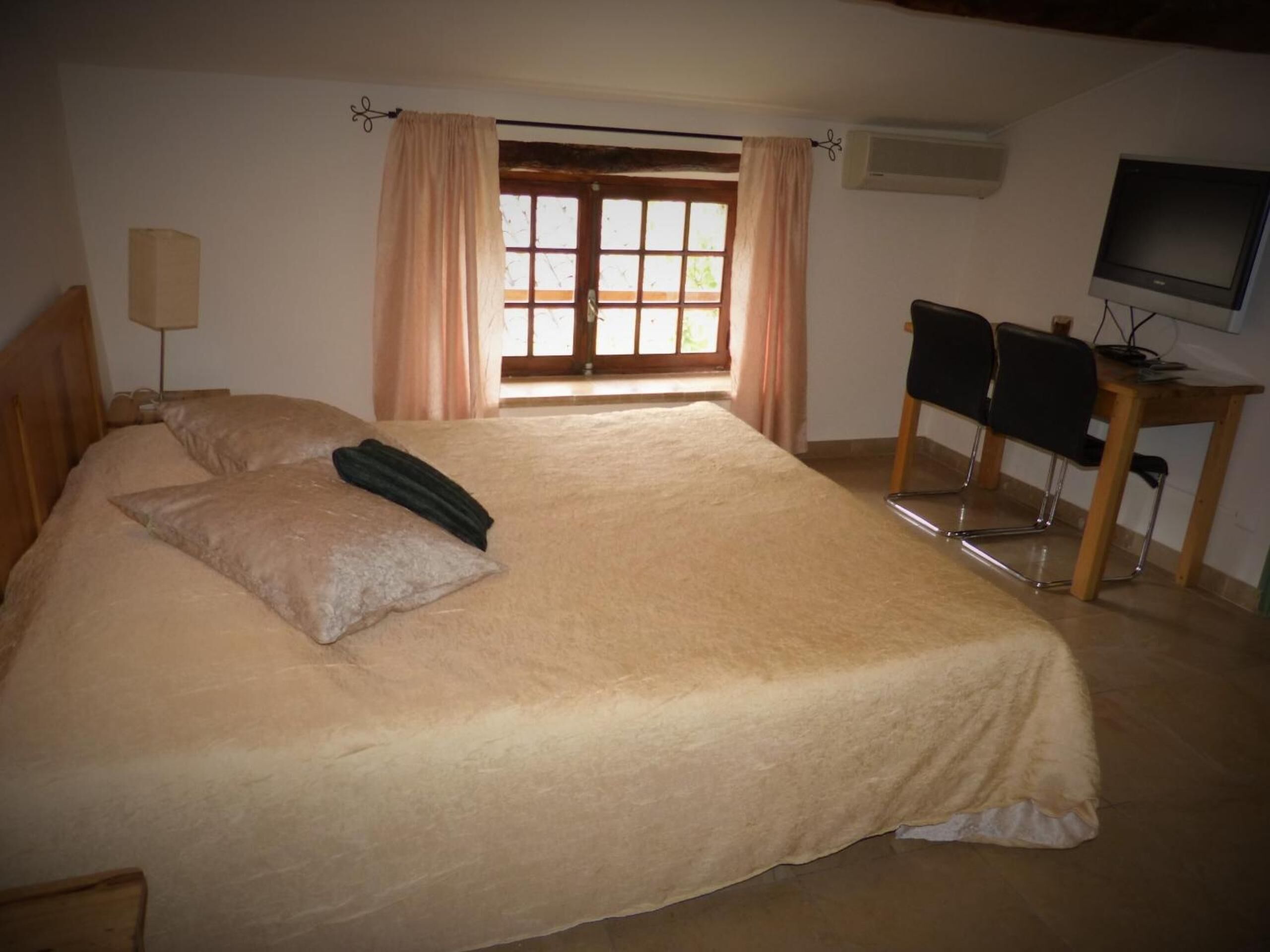 A bedroom with a large bed, a desk with a chair, a television, and a window with curtains.