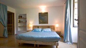 Une chambre avec un grand lit, deux tables de chevet, une commode, une lampe fixée au mur, un tableau et une porte avec des rideaux.