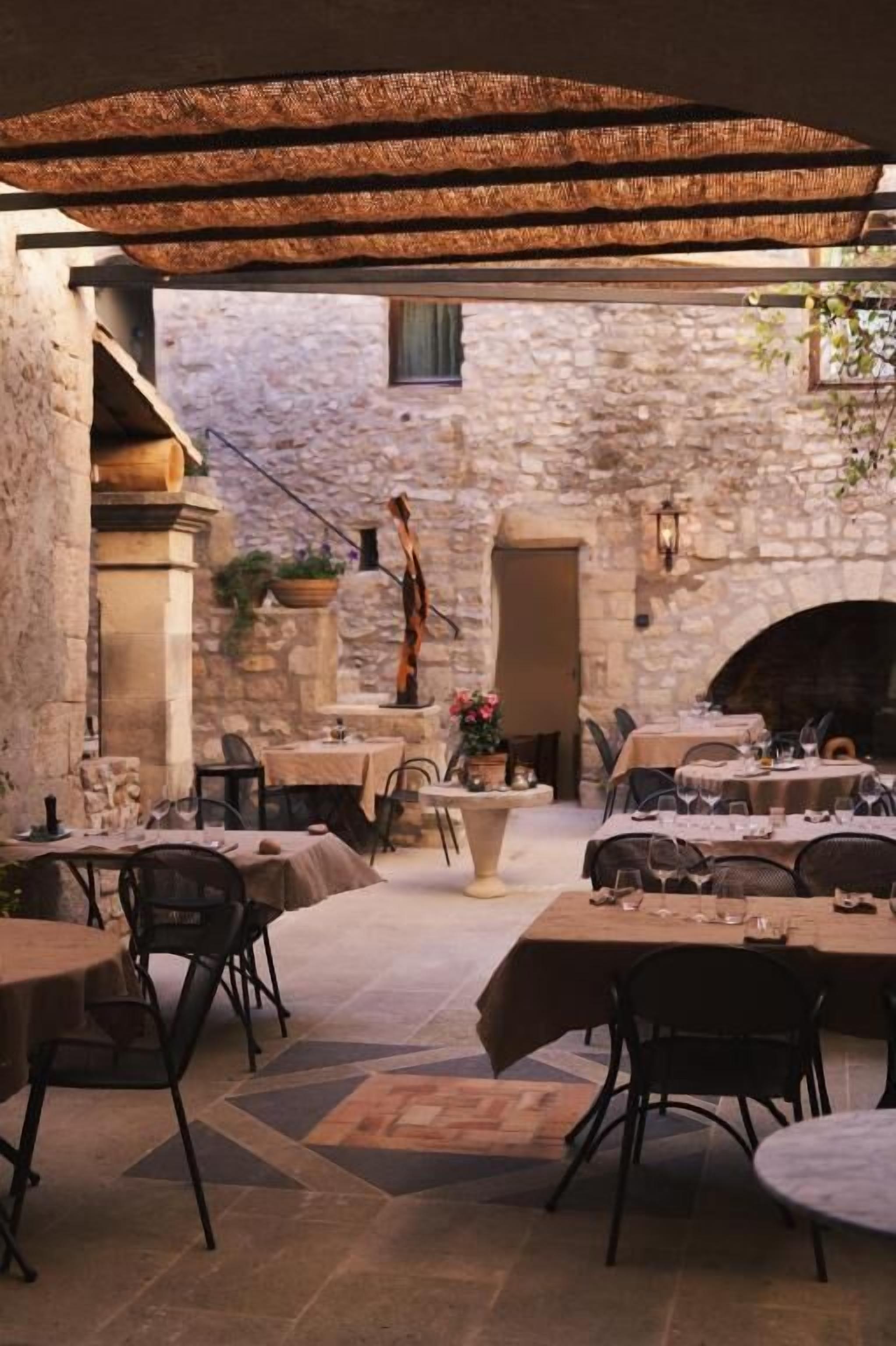 Un área de comedor al aire libre con muros de piedra, mesas, sillas y plantas colgantes.