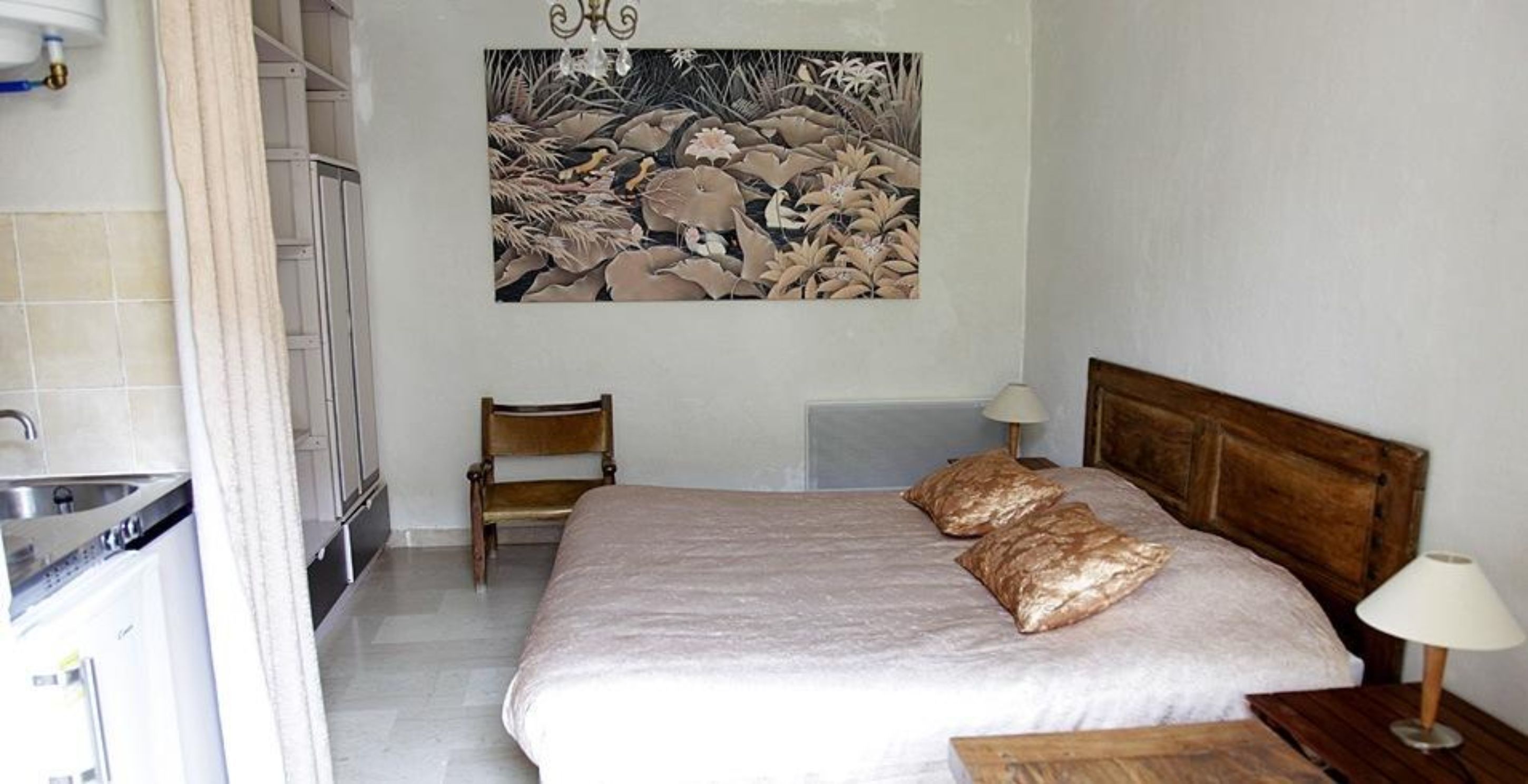 Un dormitorio con una cama, dos almohadas, una mesita de noche, una lámpara, una silla y una obra de arte colgada en la pared.