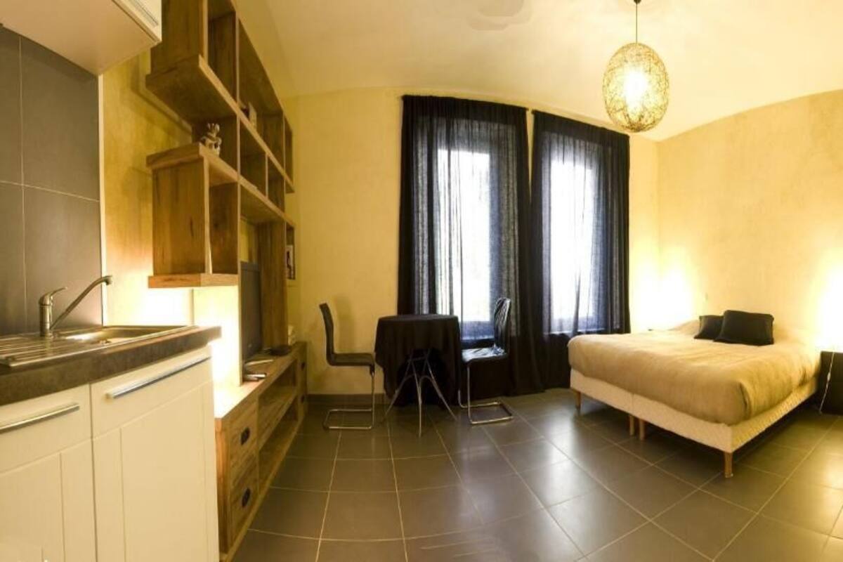 Une chambre compacte comprenant un lit, une table de chevet et un petit bureau avec une chaise. On y trouve une fenêtre avec des rideaux, une étagère intégrée et un coin cuisine avec un évier et des placards.