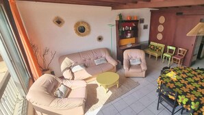 A living room with a sofa, armchairs, a coffee table, a dining table with a floral tablecloth, and a bookshelf.