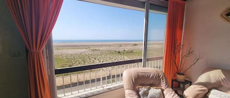 A room with a view of a beach, a chair, and a small table with a vase.