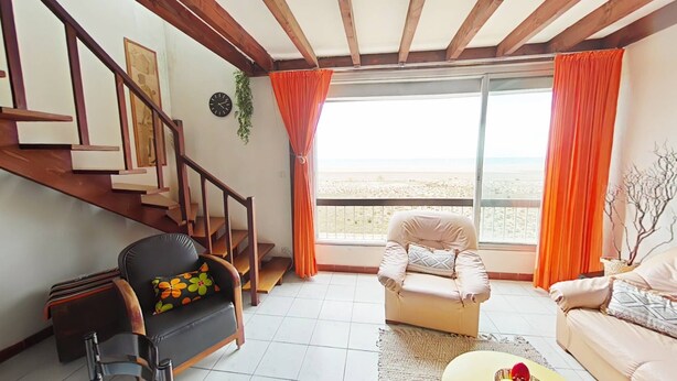 A living room with a staircase, a sofa, a chair, and a view of the beach.