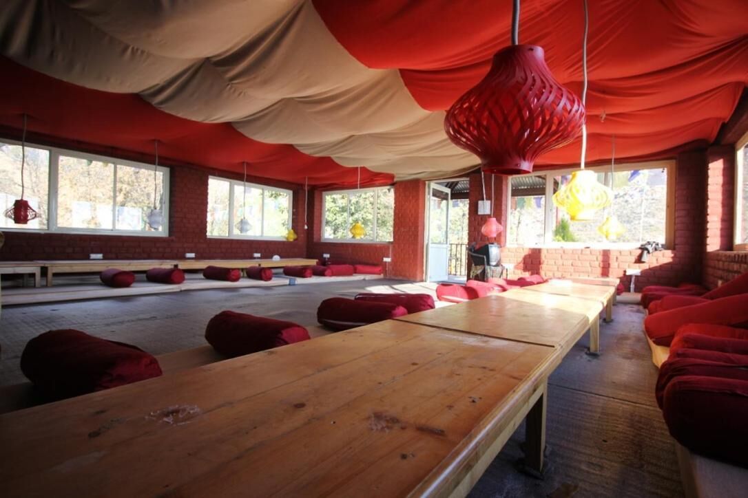 Una habitación con largas mesas de madera, asientos con cojines rojos, ventanas grandes y una lámpara colgante roja.