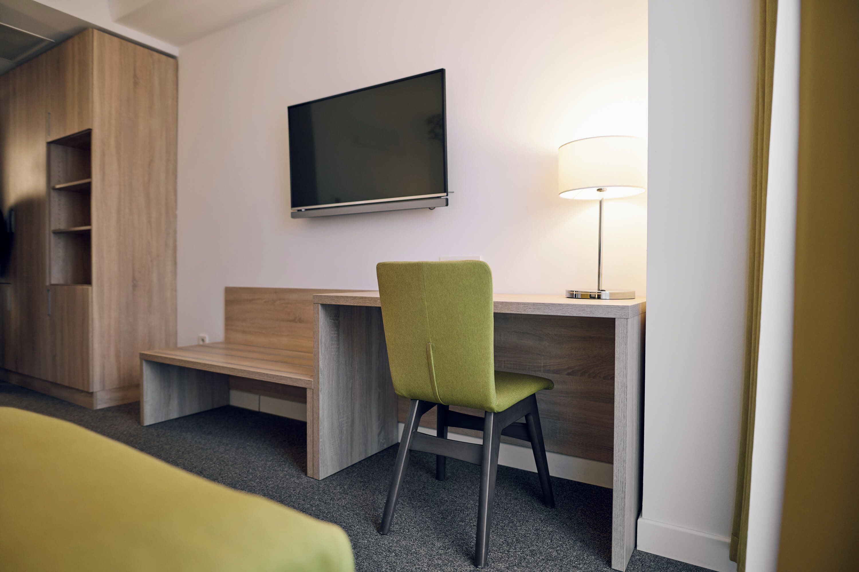 Une chambre d’hôtel avec un bureau en bois, une chaise verte, un téléviseur à écran plat et une lampe de chevet.