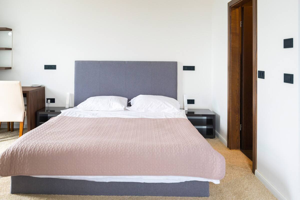 Un lit bien fait, avec une tête de lit grise, des oreillers blancs et une couverture rose. On y trouve deux tables de chevet avec des lampes, une table de nuit en bois et une porte avec un cadre en bois.