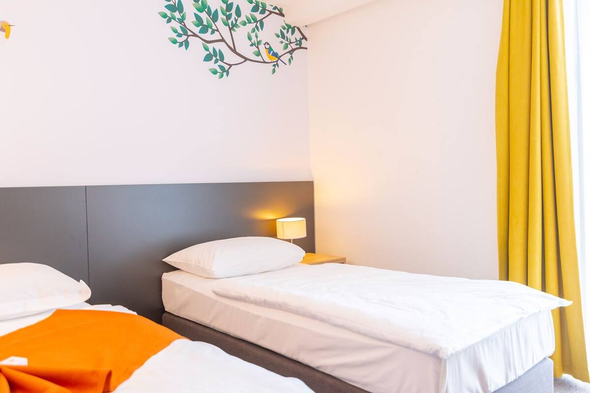 Une chambre d’hôtel avec deux lits, un motif décoratif représentant un arbre appliqué sur le mur, une lampe de chevet et une fenêtre avec des rideaux jaunes.