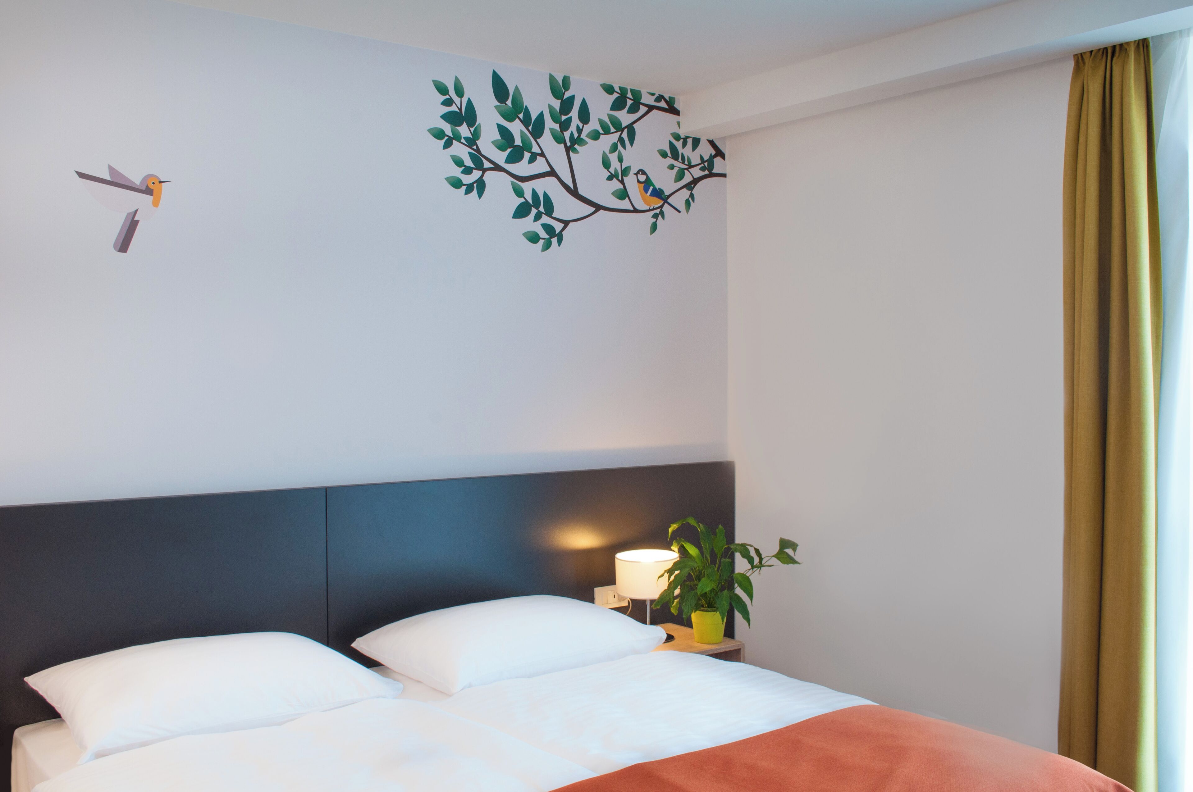 A hotel room with a bed, a nightstand with a lamp and a plant, a wall mural of a tree with birds, and a window with curtains.