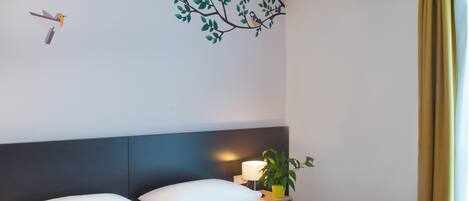A hotel room with a bed, a nightstand with a lamp and a plant, a wall mural of a tree with birds, and a window with curtains.