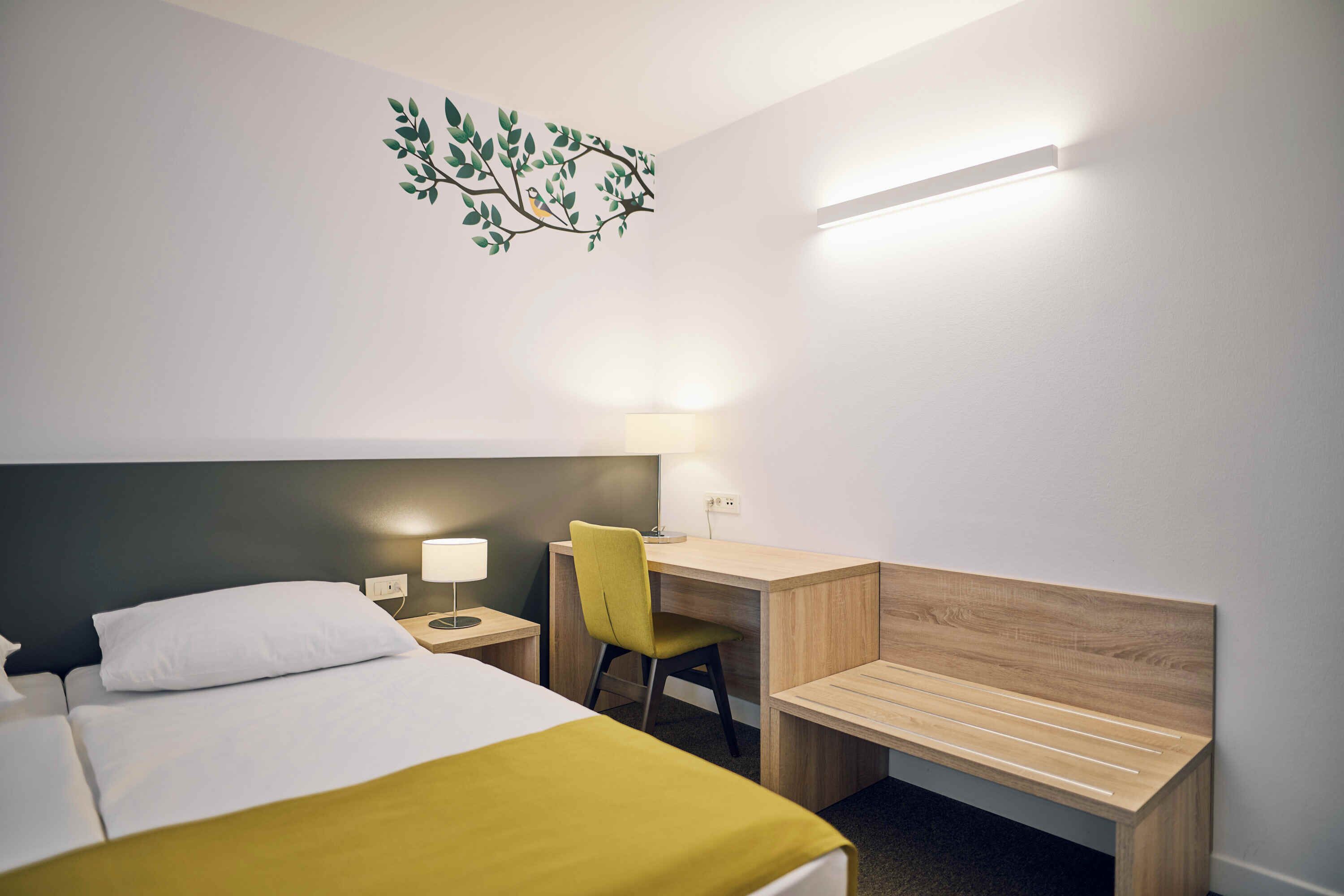 A hotel room with a bed, a desk, a chair, and a wall-mounted lamp.