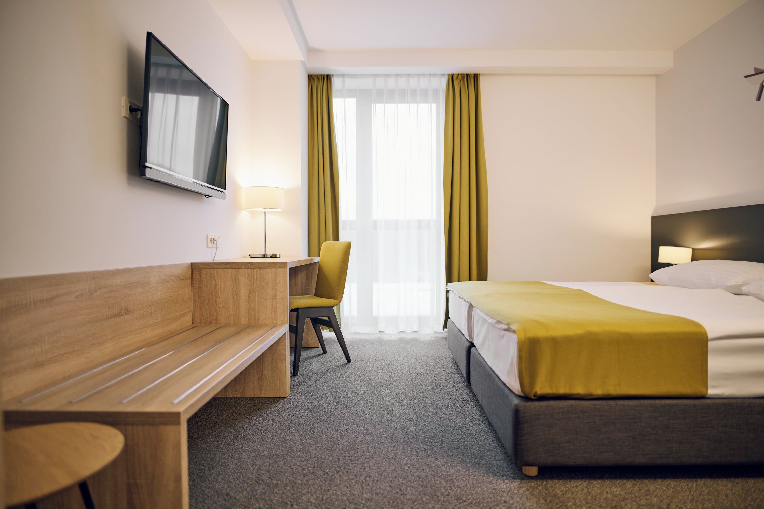 A hotel room with a bed, a desk, a chair, a TV mounted on the wall, and a window with curtains.