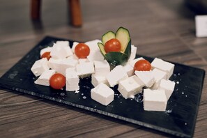 Un plateau de service noir en ardoise, sur lequel on trouve des cubes de fromage blanc, des tomates cerises et une tranche de concombre, le tout posé sur un plancher en bois.