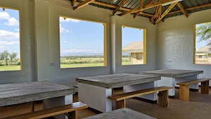 Una habitación con mesas y bancos de concreto, ventanas amplias con vistas a un paisaje verde y un techo de lámina corrugada.