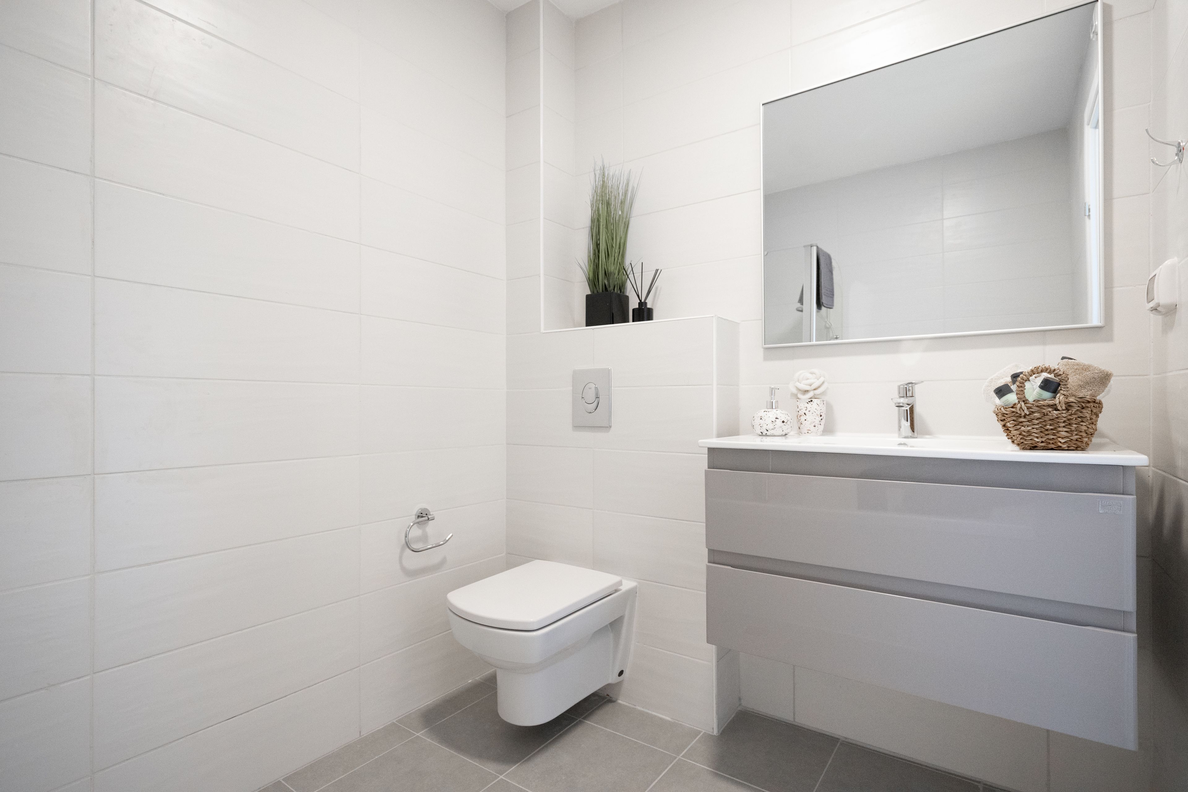 A modern bathroom with white tiles, a toilet, a sink with a mirror, and a basket on the counter.