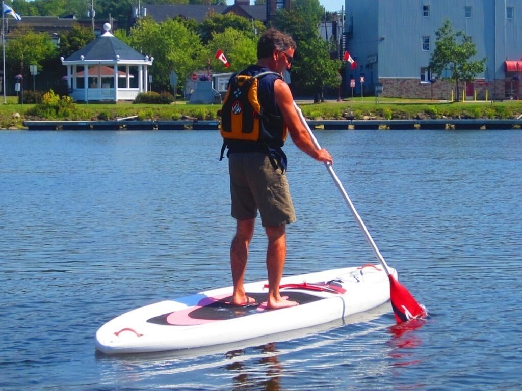 Una persona de pie en una tabla de remo en un cuerpo de agua, con un pabellón y edificios al fondo.