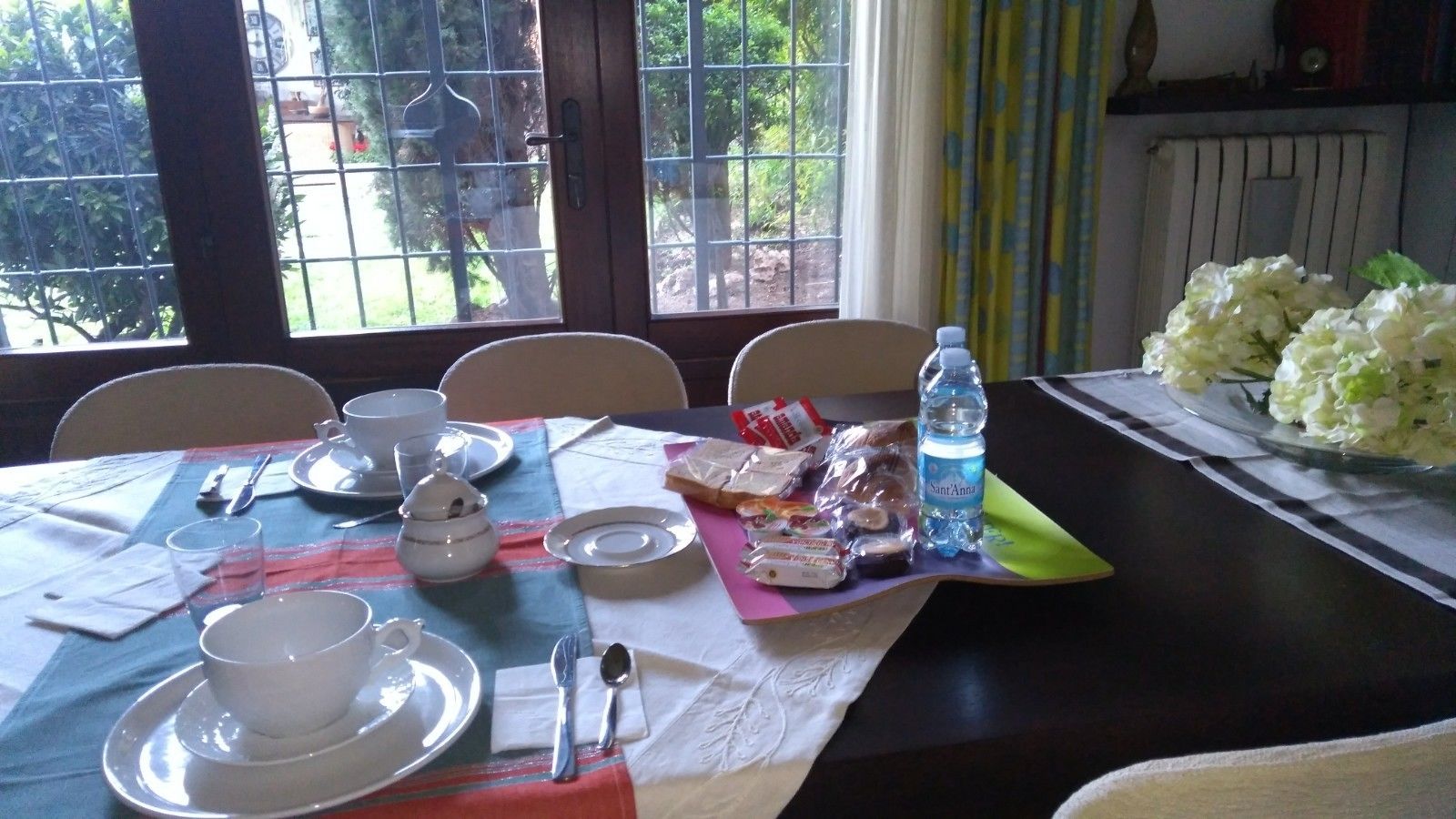 Une table à manger dressée avec de l’assiette blanche, des tasses et des couverts, une bouteille d’eau et des emballages alimentaires.